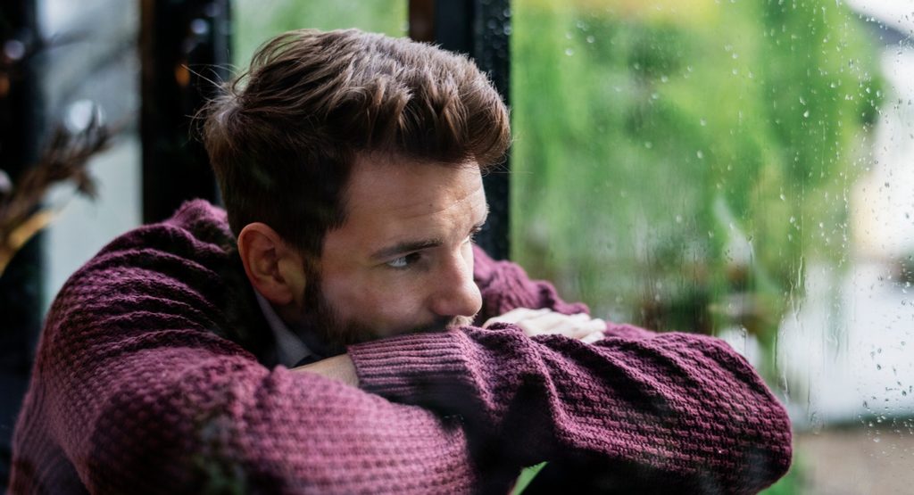 Man looking out a rainy window, reflecting the emotional challenges of depression and mental health struggles.